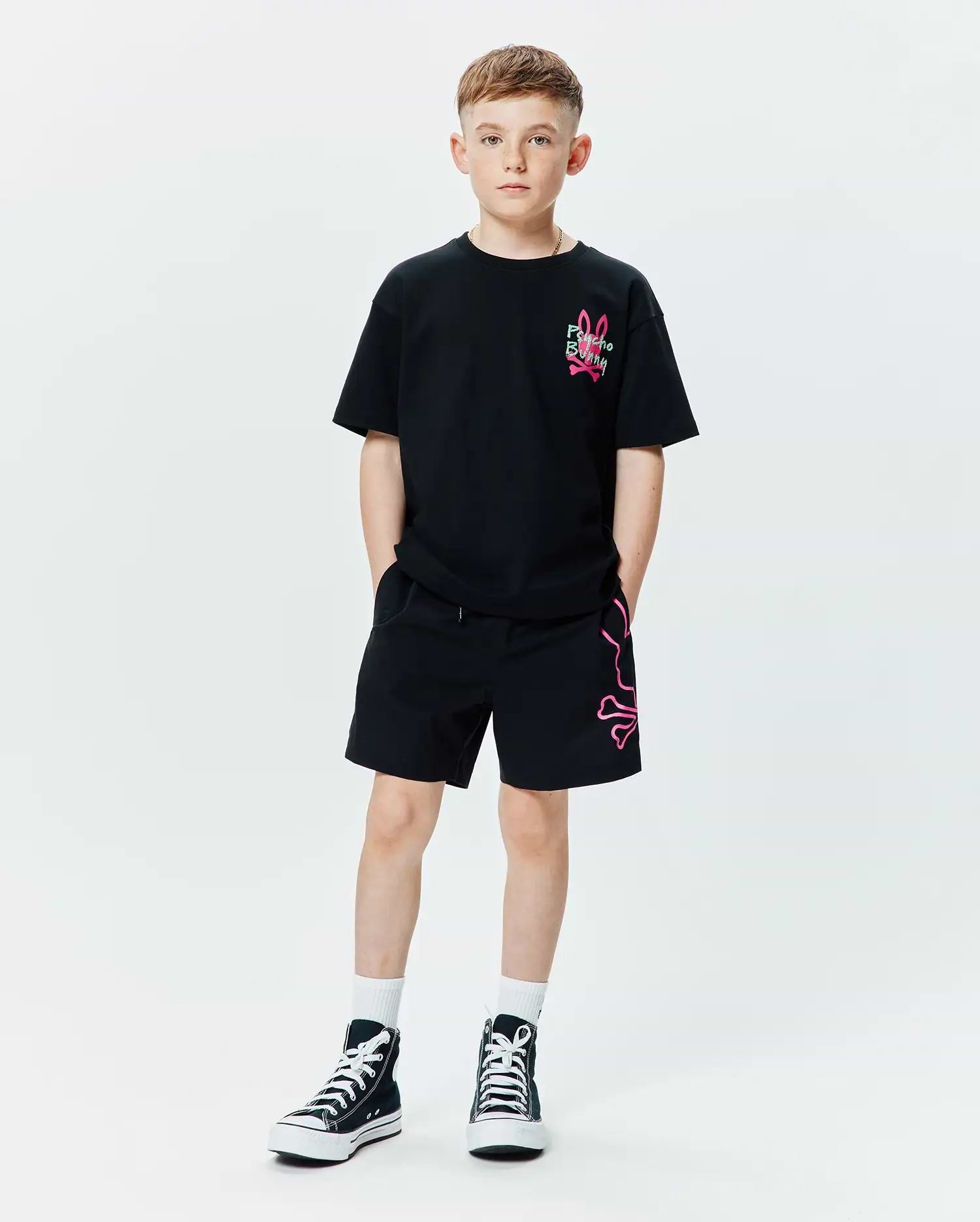 Kids Duncan Relaxed Fit Tee in black jersey with a small neon pink embroidered Psycho Bunny logo on the chest, paired with black relaxed fit shorts featuring a large neon pink outline bunny skull graphic on the left leg.