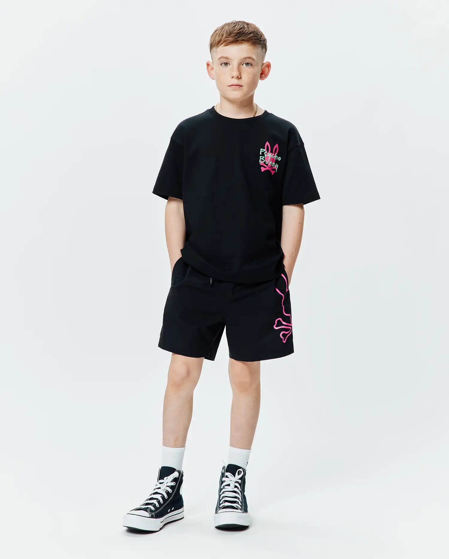 Kids Duncan Relaxed Fit Tee in black jersey with a small neon pink embroidered Psycho Bunny logo on the chest, paired with black relaxed fit shorts featuring a large neon pink outline bunny skull graphic on the left leg.