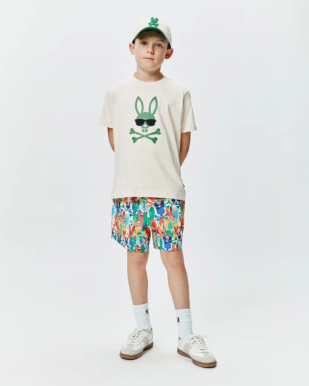 Ecru jersey Kids Sunny Graphic Tee featuring a large green Psycho Bunny skull graphic with sunglasses, complemented by multicolor all-over bunny print shorts and a white embroidered logo cap.