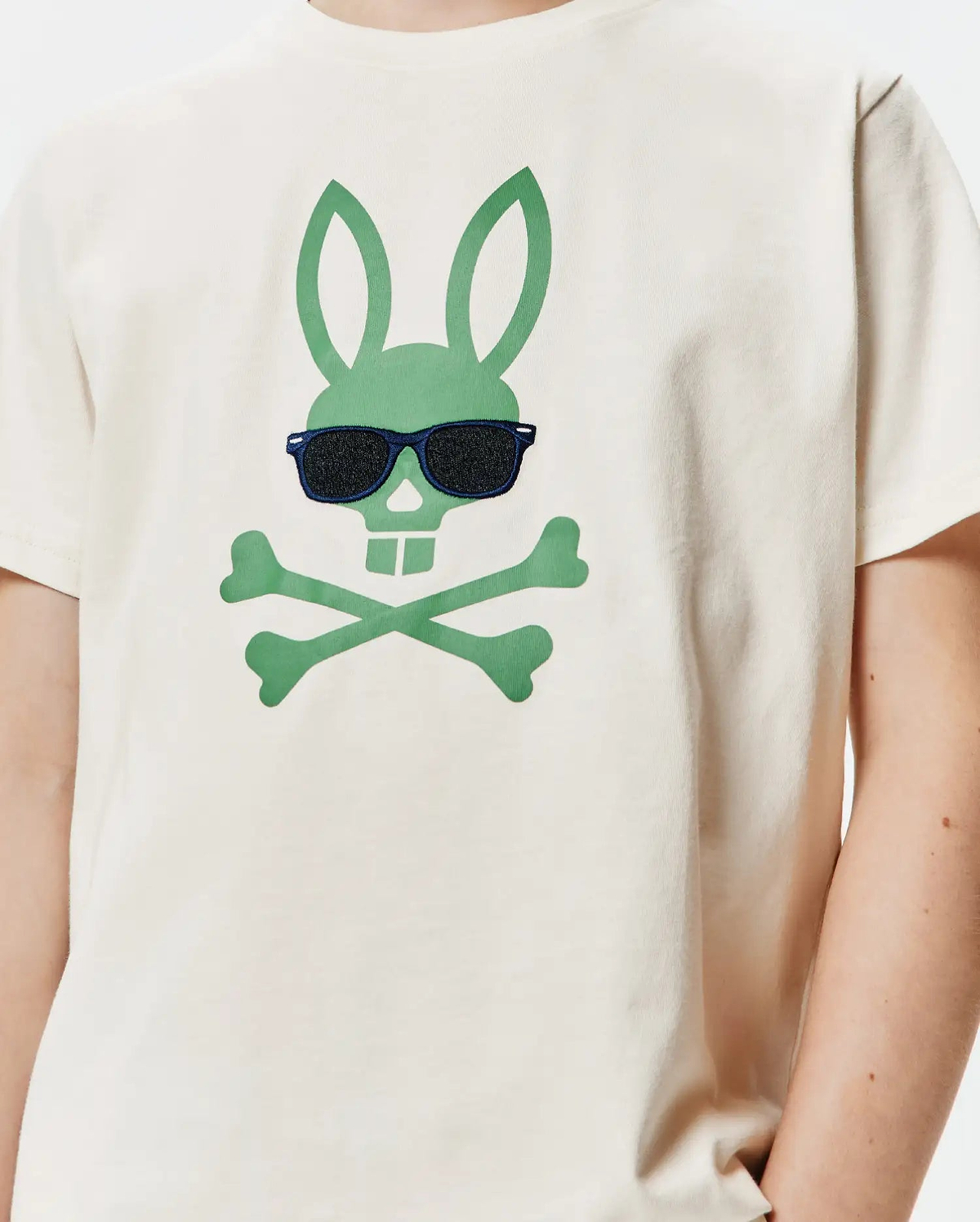 Kids Sunny Graphic Tee, a cream classic-fit jersey t-shirt featuring a large central green Psycho Bunny skull and crossbones logo wearing embroidered dark blue sunglasses with black lenses.