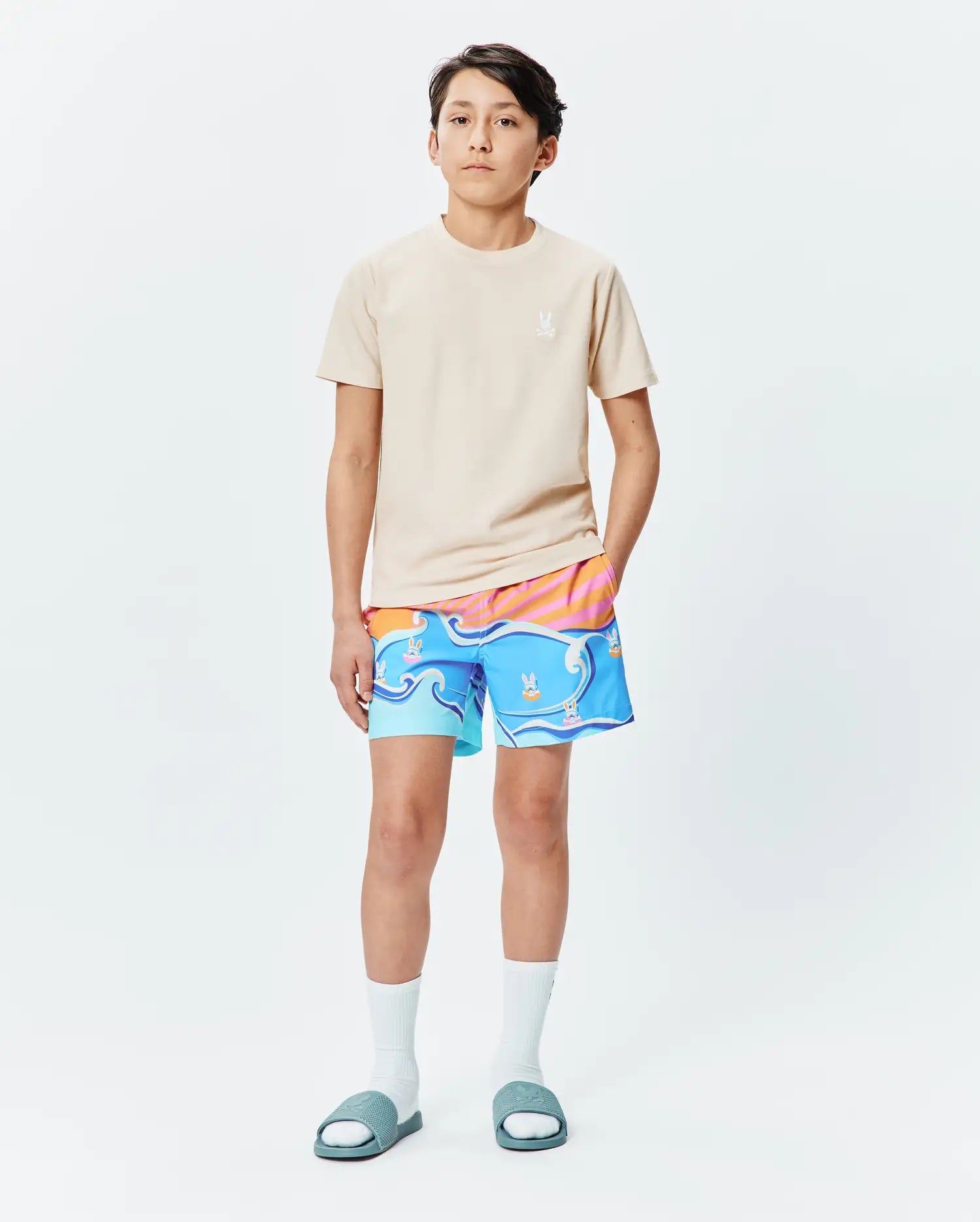 Kids Taylor Pique Tee in light tan, classic fit, with a small embroidered white Psycho Bunny skull logo on the left chest, paired with vibrant blue and orange surf-patterned swim shorts featuring bunny skull graphics.