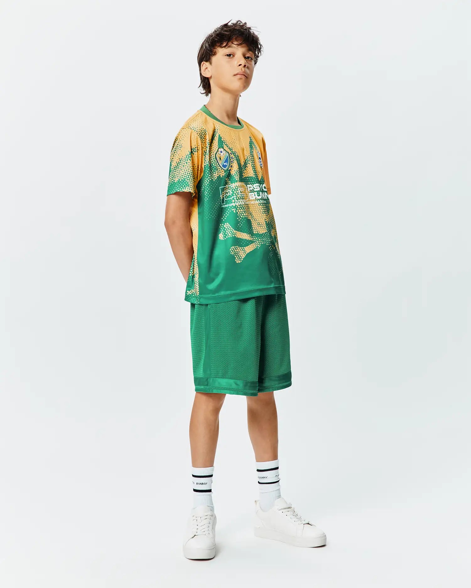 Kids Joshua Soccer Tee and matching green mesh shorts. The tee features a vibrant gold to green gradient with a distressed skull pattern, a small bunny skull crest, and large white Psycho Bunny text. Relaxed fit.