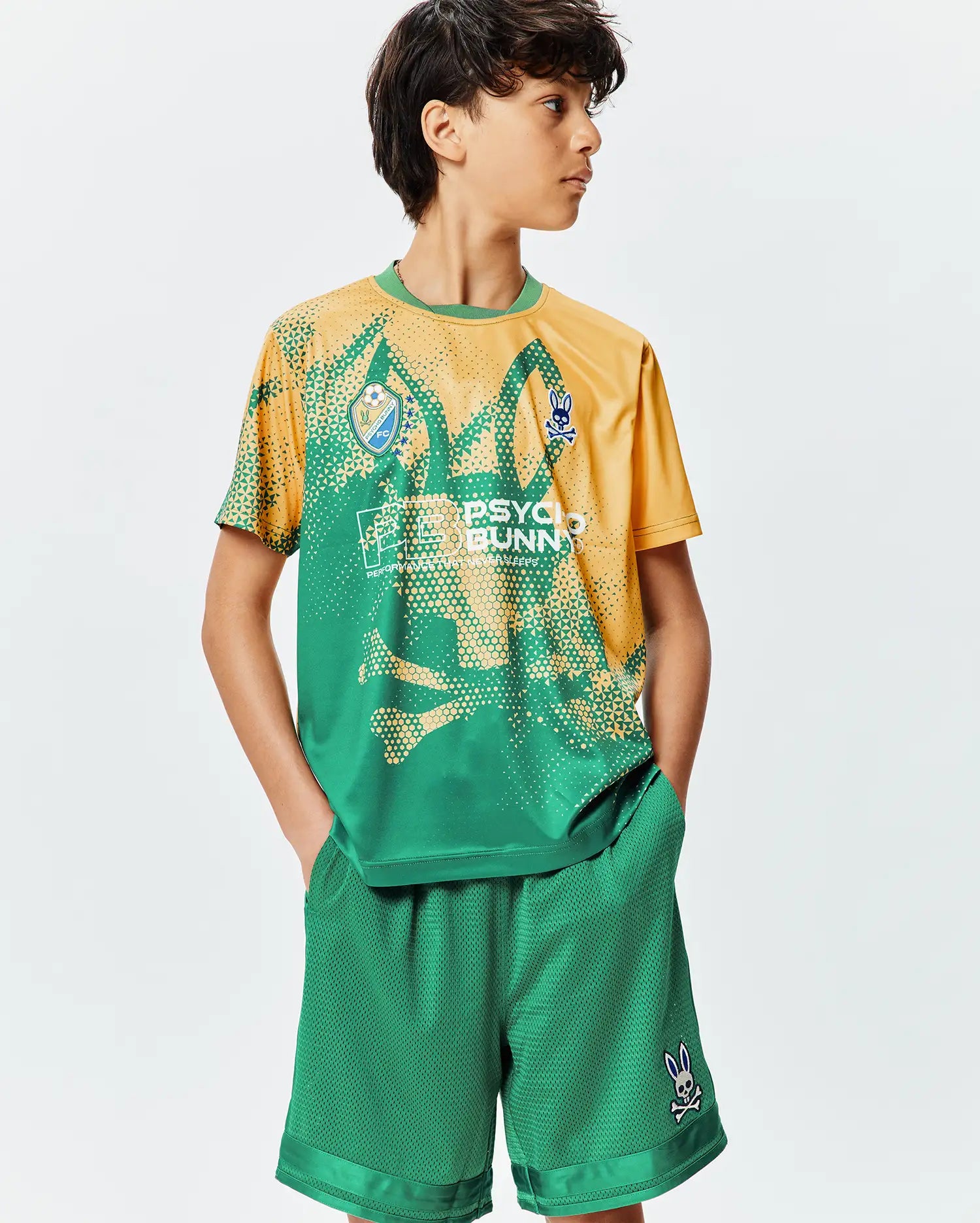 Kids Joshua Soccer Tee and green mesh shorts. The relaxed fit tee has a green to gold gradient, pixelated pattern, bold PSYCHO BUNNY text, FC logo, and small bunny skull logos. Shorts feature a small bunny skull.