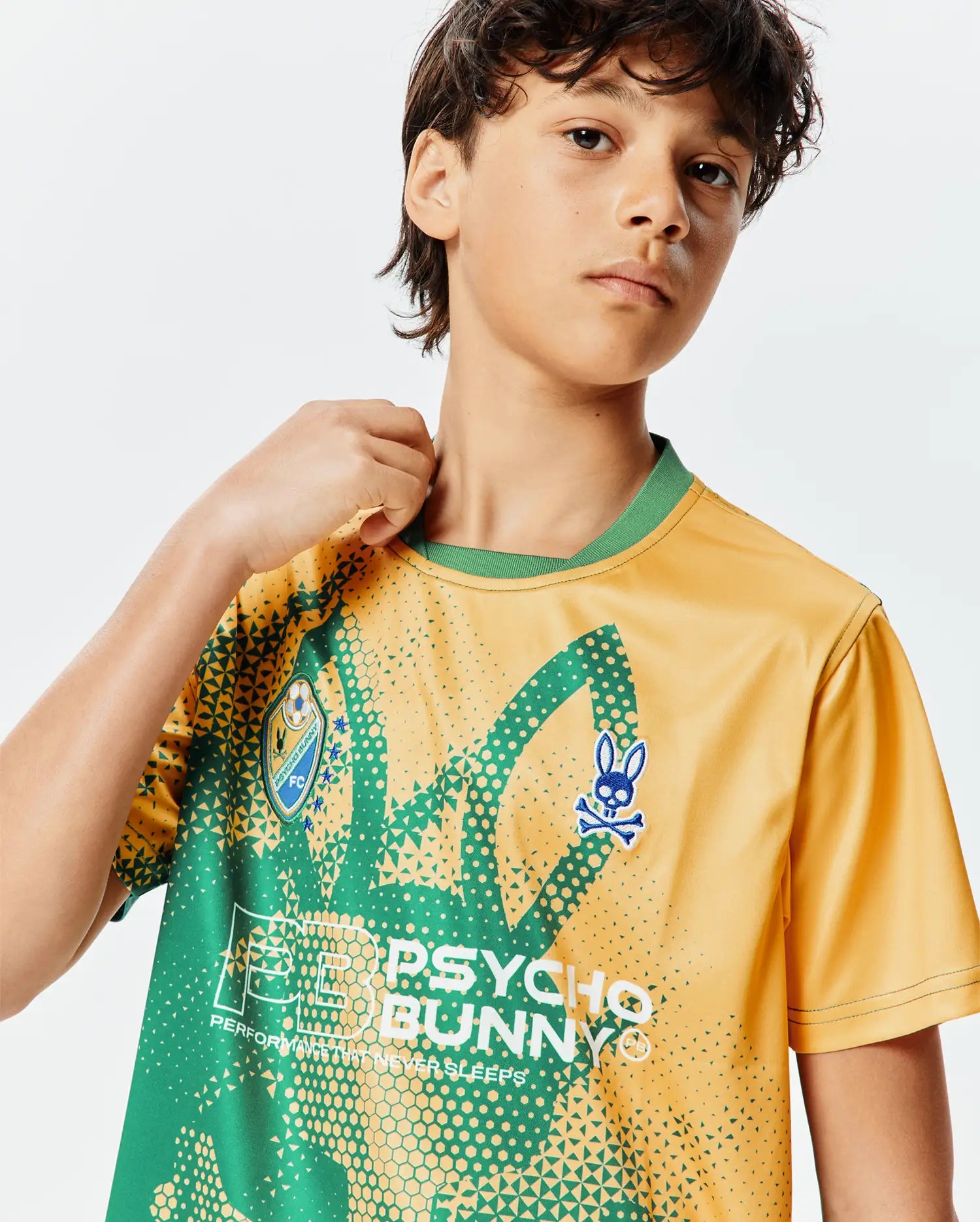 Kids Joshua Soccer Tee in gold with a vibrant green geometric pattern, featuring a large Psycho Bunny text graphic, an embroidered blue bunny skull logo, and a soccer club patch. Classic fit, smooth jersey.