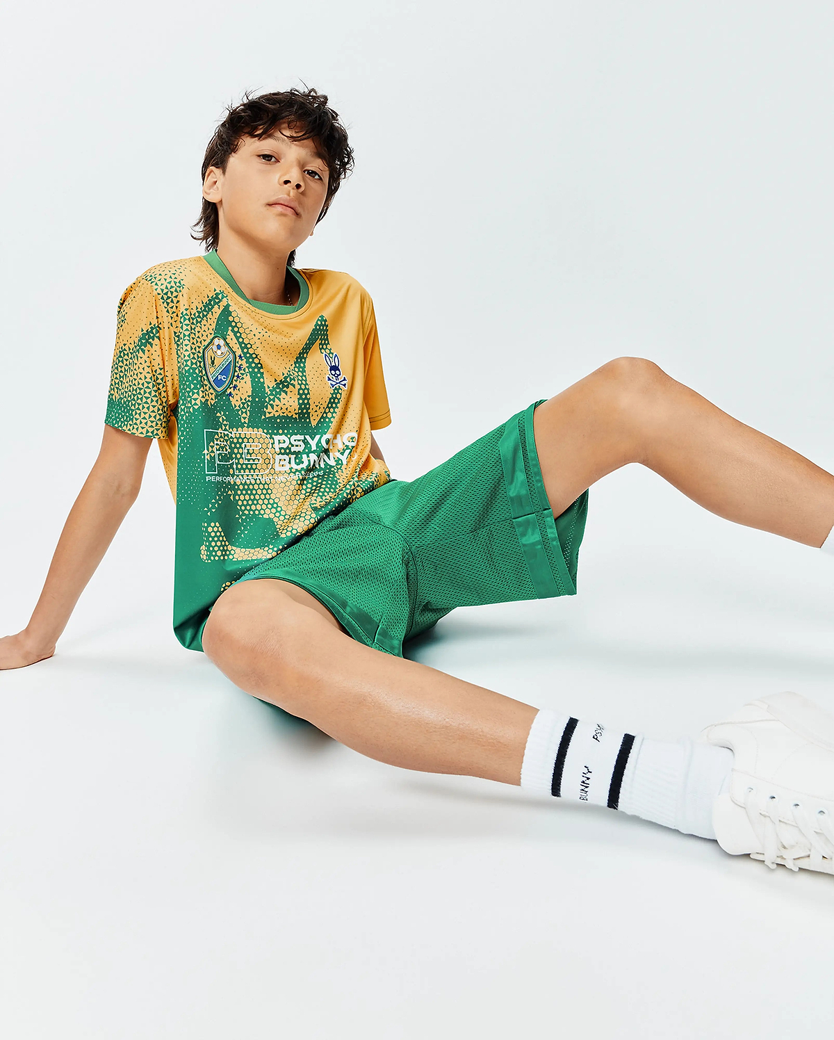Kids Joshua Soccer Tee, a relaxed fit yellow and green performance jersey with geometric patterns, large Psycho Bunny text, and small bunny skull and FC shield logos on the chest, paired with green mesh shorts.