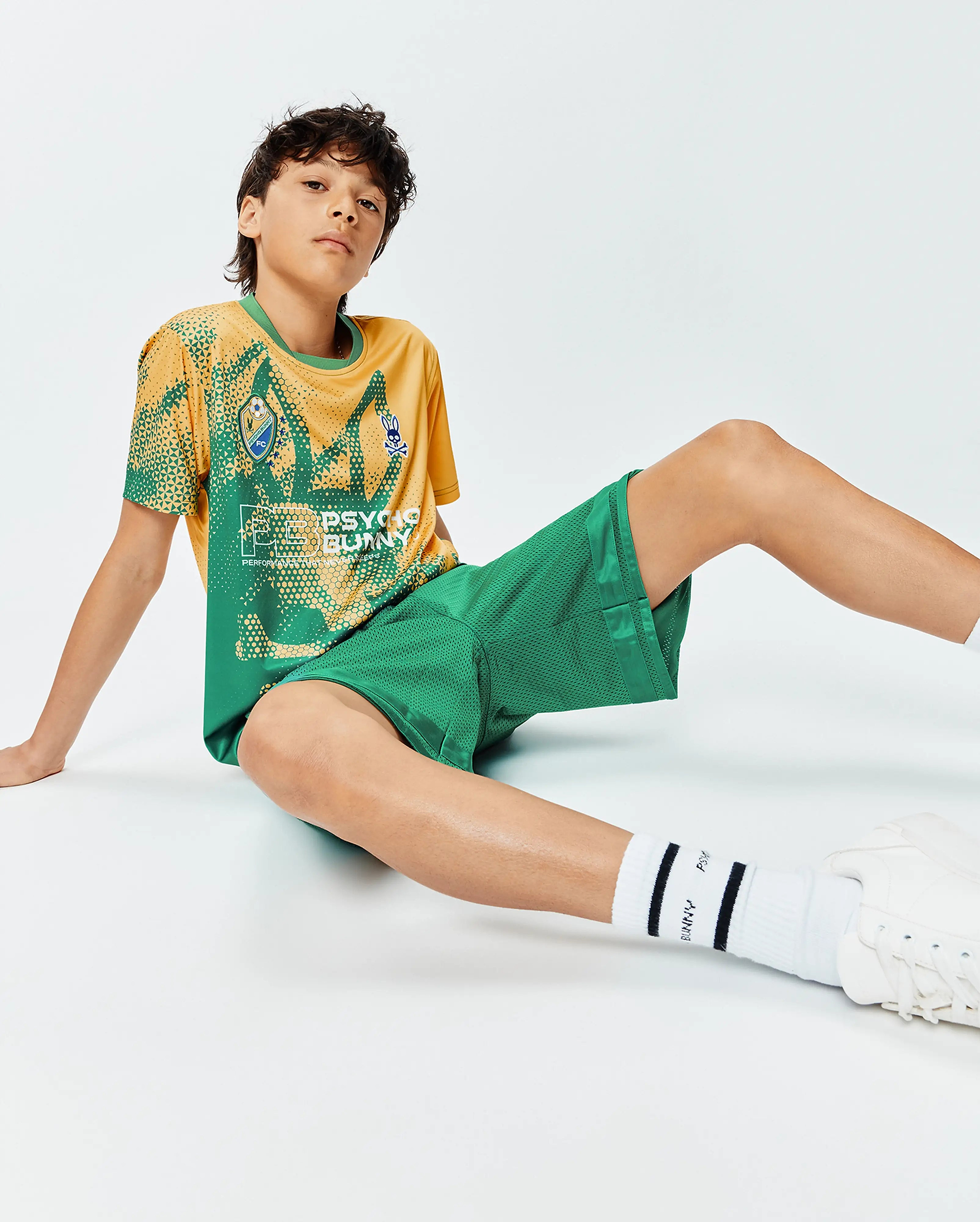 Kids Joshua Soccer Tee, a relaxed fit yellow and green performance jersey with geometric patterns, large Psycho Bunny text, and small bunny skull and FC shield logos on the chest, paired with green mesh shorts.