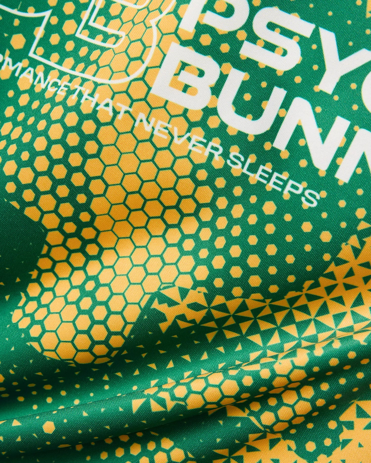 Kids Joshua Soccer Tee in vibrant green performance jersey, featuring bold yellow geometric hexagonal and triangular patterns, with large white PSYCHO BUNNY text and a PERFORMANCE THAT NEVER SLEEPS tagline.