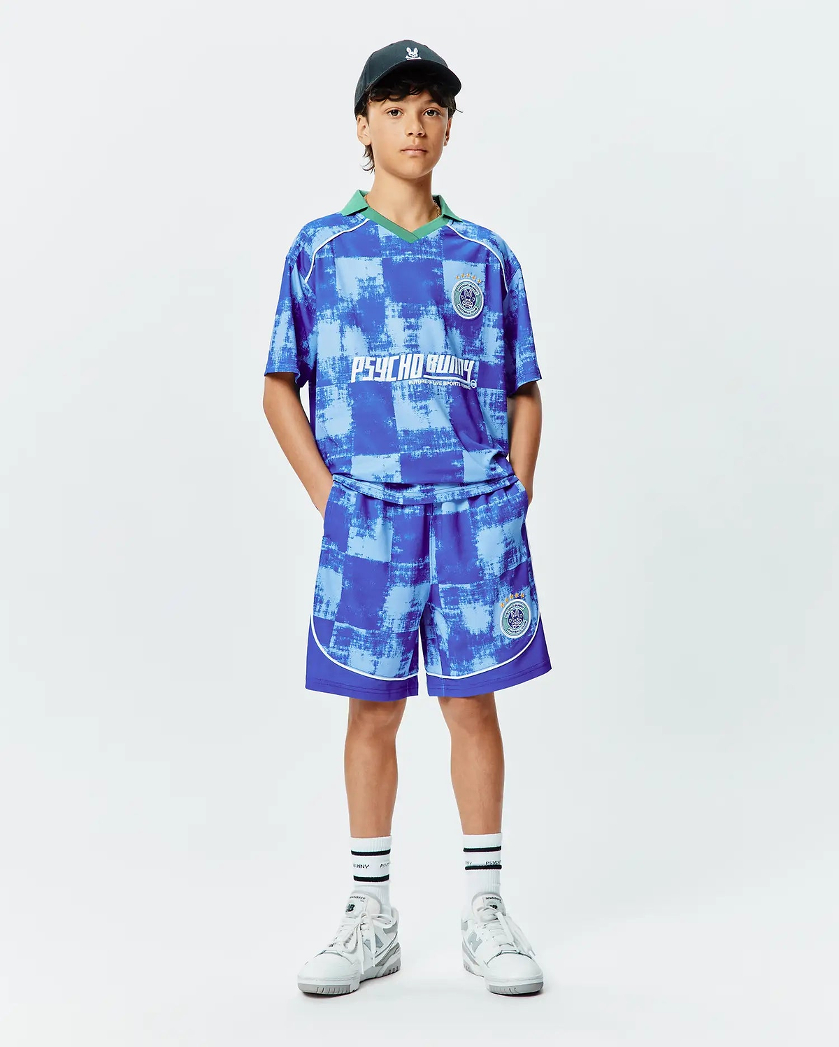 KIDS MICAH OVERSIZED SOCCER JERSEY and matching shorts in a blue distressed checkered pattern. Features a green V-neck, white PSYCHO BUNNY chest graphic, and circular crest logos. White piping details.