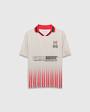 Beige Kids Camden Oversized Soccer Jersey with a red and black striped collar, embroidered Psycho Bunny skull logo on the left chest, large PSYCHO BUNNY graphic, and fading red vertical stripes on the front.