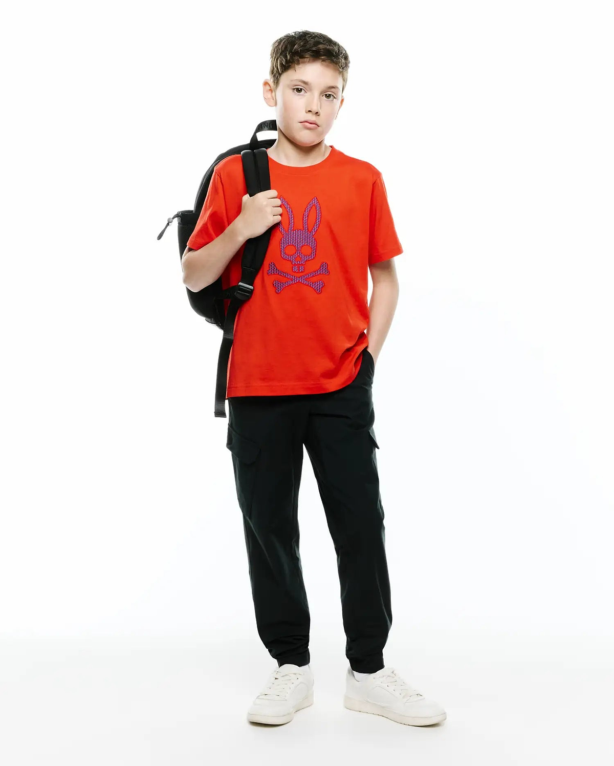 Vibrant orange Kids Stan Graphic Tee, a classic fit jersey t-shirt featuring a large purple and blue Psycho Bunny skull logo on the chest, paired with relaxed dark green cargo pants.