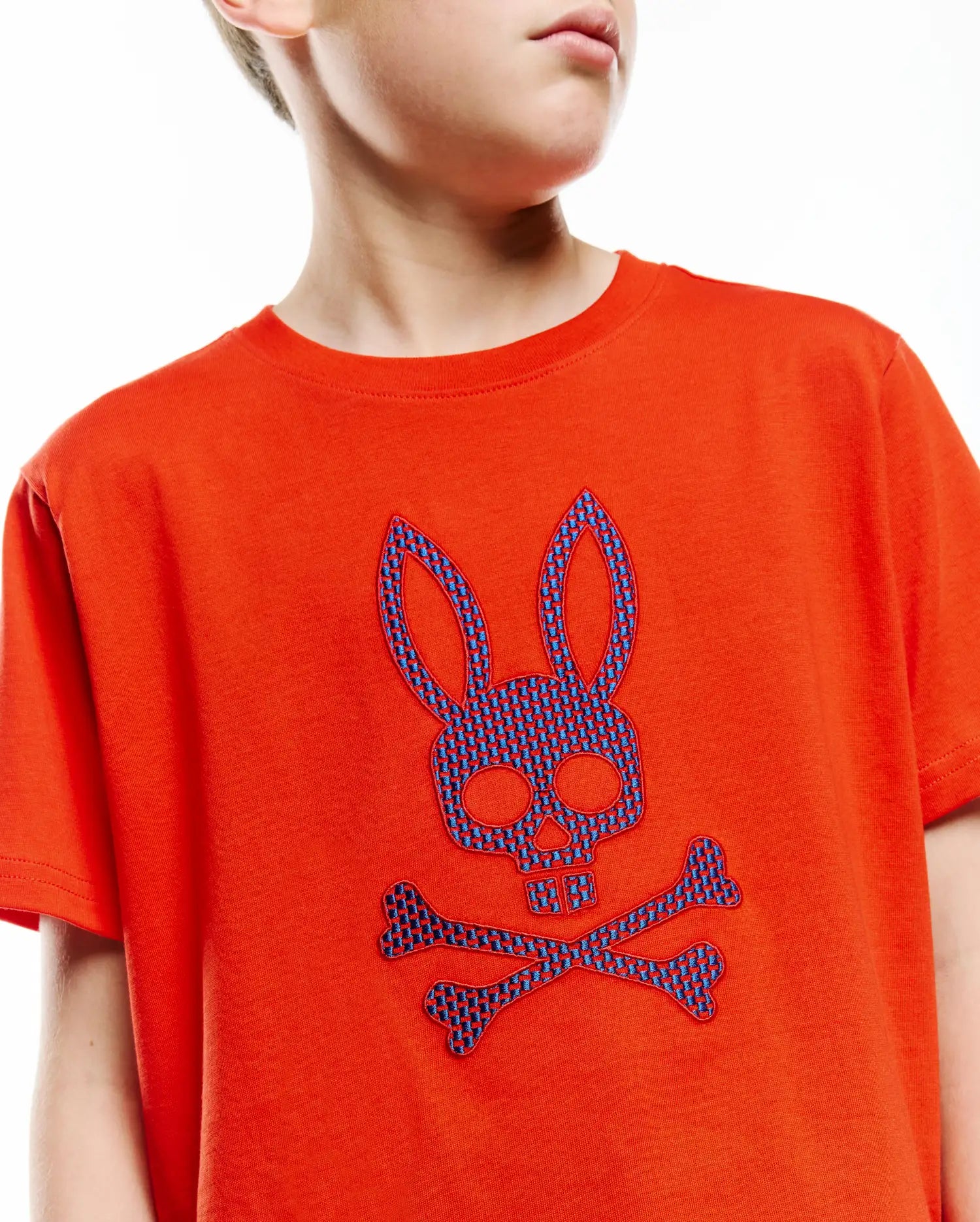 Kids Stan Graphic Tee, vibrant orange jersey knit with a large, central embroidered blue checkered Psycho Bunny skull and crossbones logo, classic fit.
