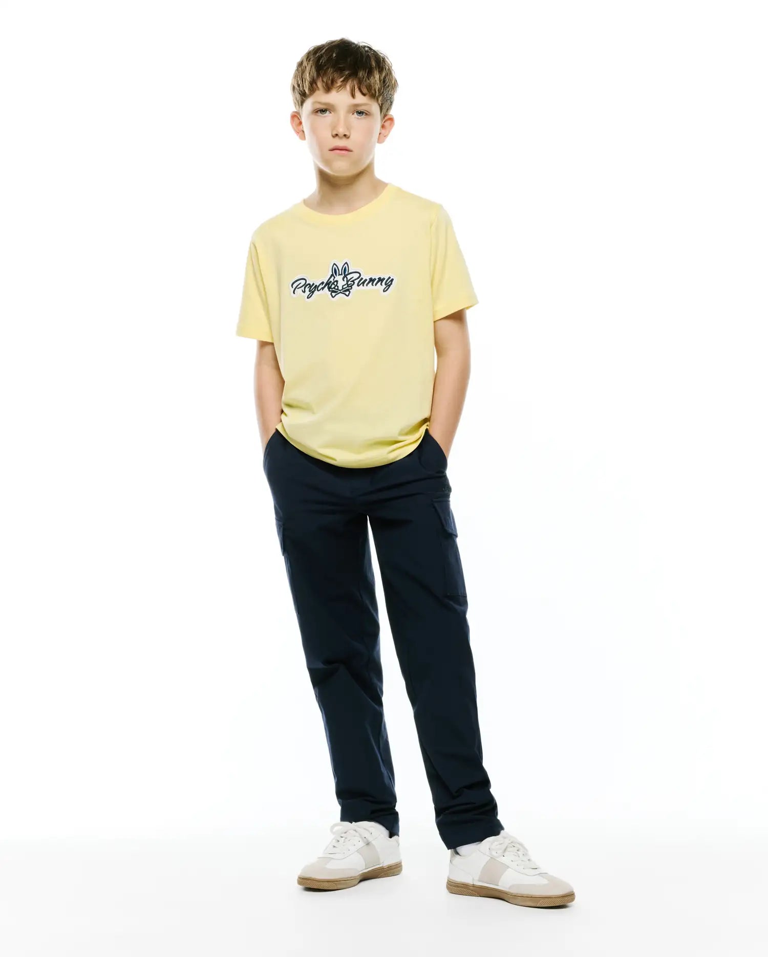 Kids Jaydon Wordmark Graphic Tee, a pale yellow classic fit jersey t-shirt with a medium blue embroidered Psycho Bunny wordmark logo on the center chest, styled with navy cargo pants.