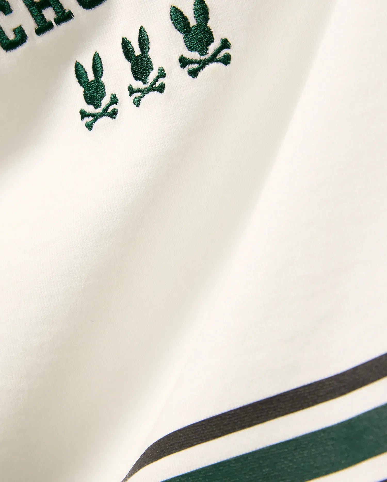 Kids Mike Graphic Jersey Tee in white jersey knit, featuring multiple small embroidered dark green Psycho Bunny skull logos and a bottom stripe design with dark green and black accents.