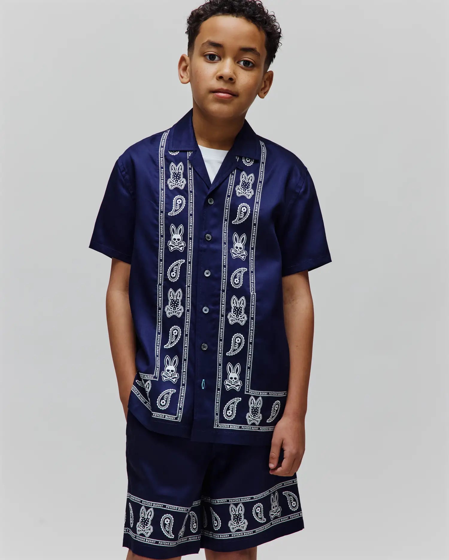 Kids Luke Tencel Border Shirt and matching shorts in navy with a white bandana-style border print featuring repeated Psycho Bunny skull logos, paisley motifs, and brand text. Relaxed fit.