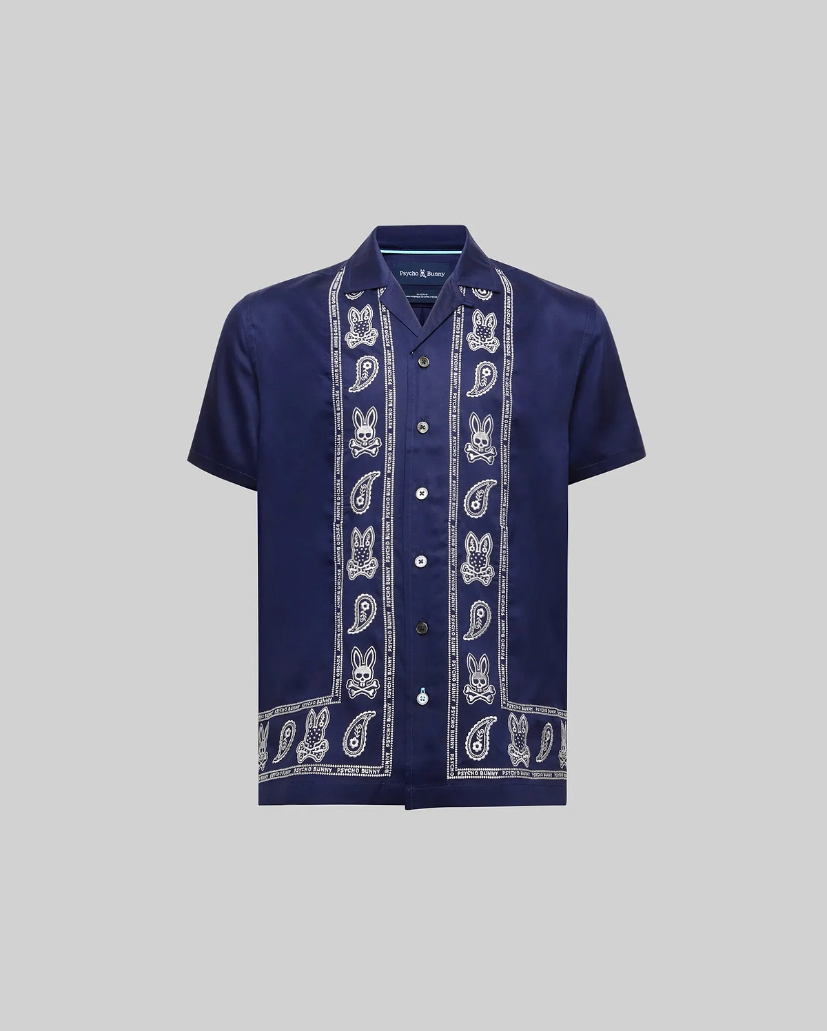Kids Luke Tencel Border Shirt, a navy blue relaxed fit short-sleeve shirt in smooth Tencel. Features white vertical Psycho Bunny skull and paisley border patterns on the placket and hem, with a small embroidered chest logo.