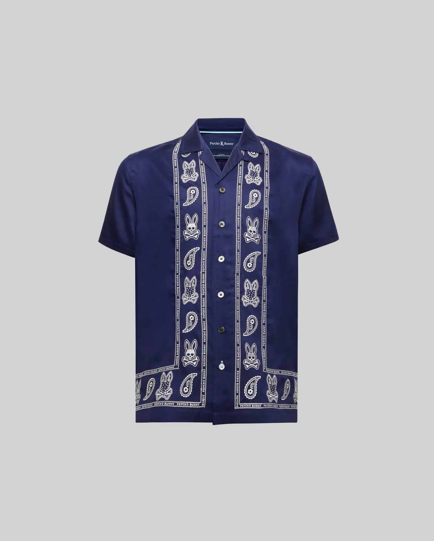 Kids Luke Tencel Border Shirt, a navy blue relaxed fit short-sleeve shirt in smooth Tencel. Features white vertical Psycho Bunny skull and paisley border patterns on the placket and hem, with a small embroidered chest logo.