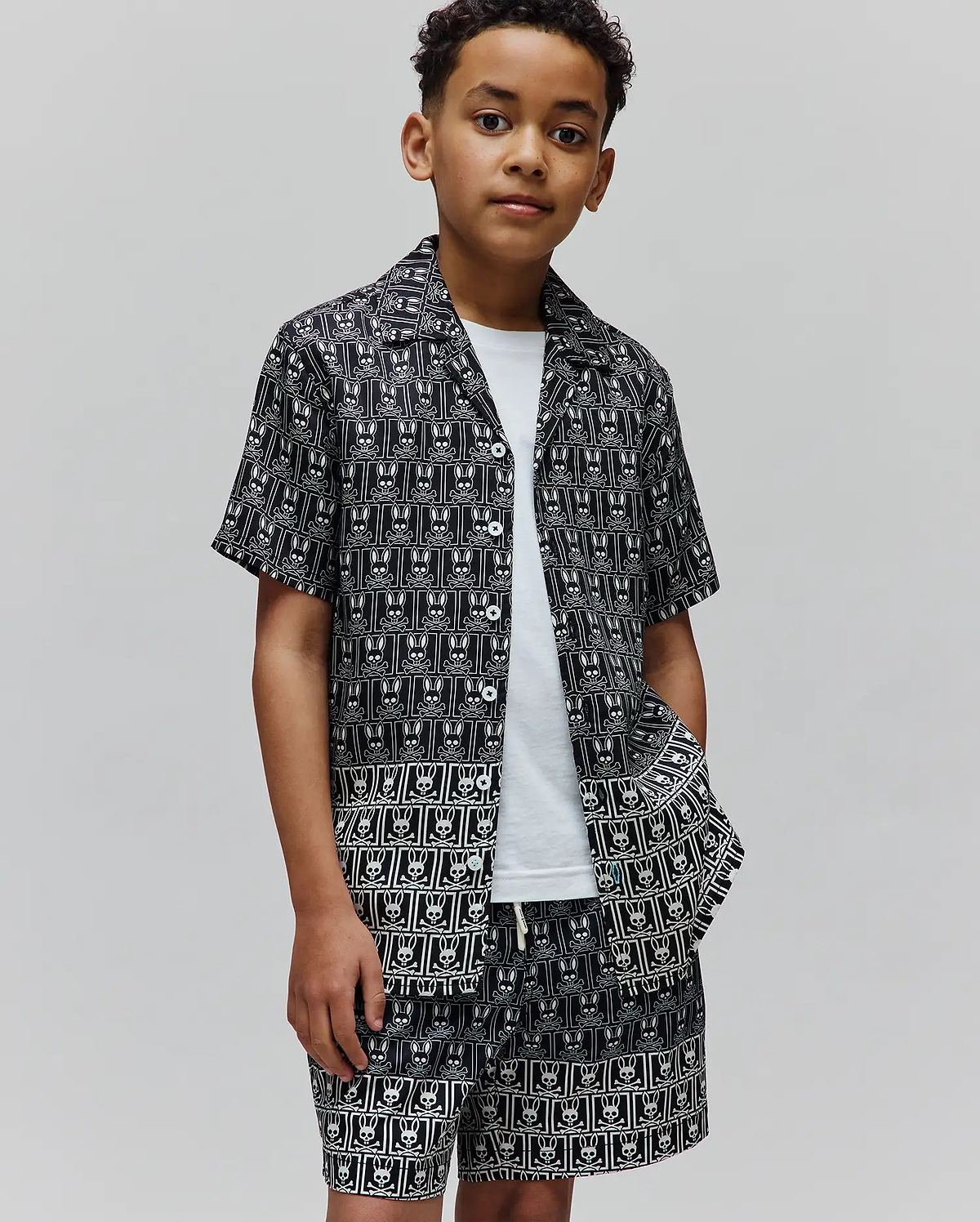 Kids Keon Tencel Shirt and matching shorts in black with an all-over white Psycho Bunny skull and crossbones pattern, featuring a relaxed fit, short sleeves, camp collar, and white buttons.