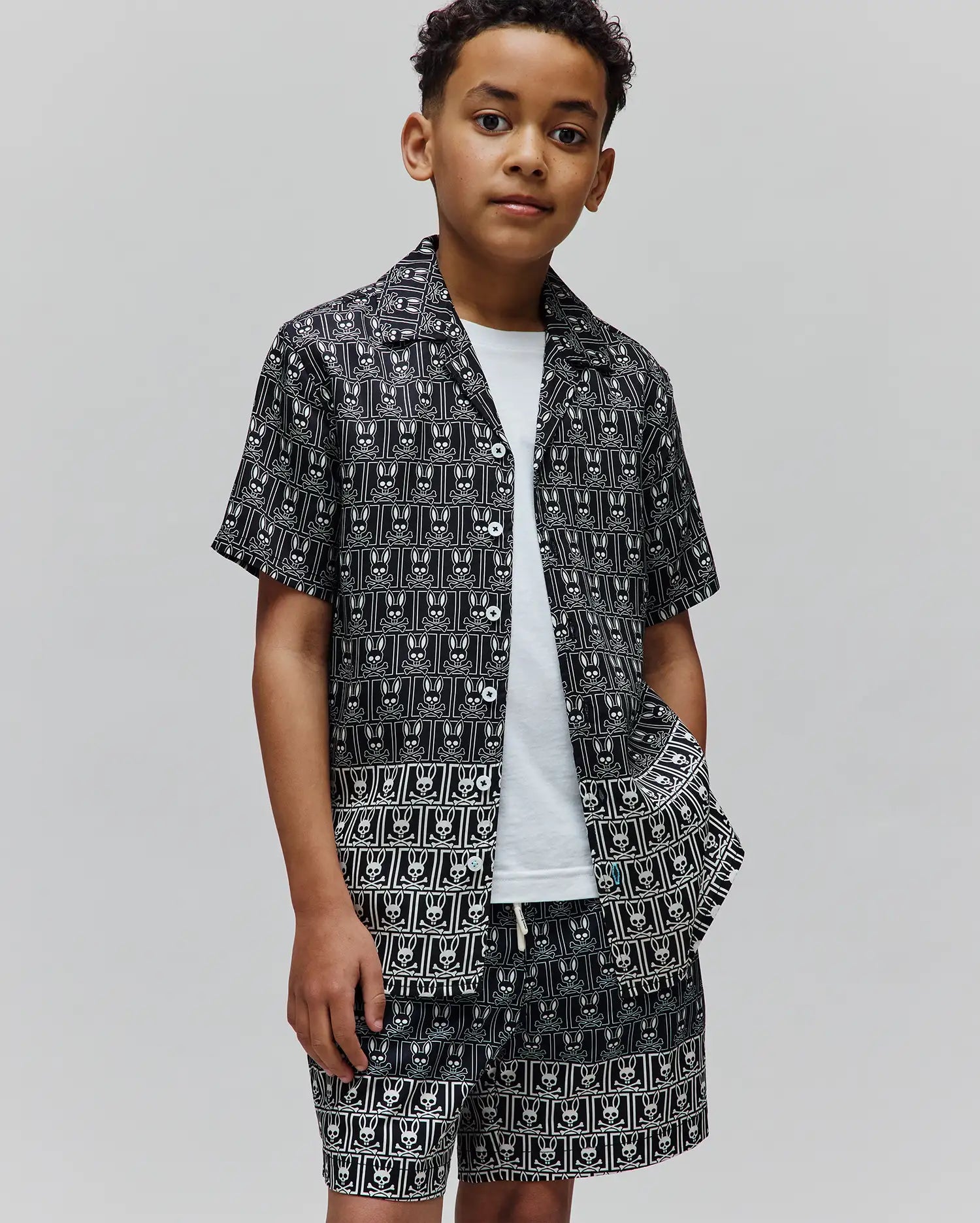 Kids Keon Tencel Shirt and matching shorts in black with an all-over white Psycho Bunny skull and crossbones pattern, featuring a relaxed fit, short sleeves, camp collar, and white buttons.