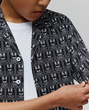 Kids Keon Tencel Shirt, a black short-sleeve woven shirt featuring a classic fit and an all-over white Psycho Bunny skull and crossbones logo pattern, complete with white buttons.