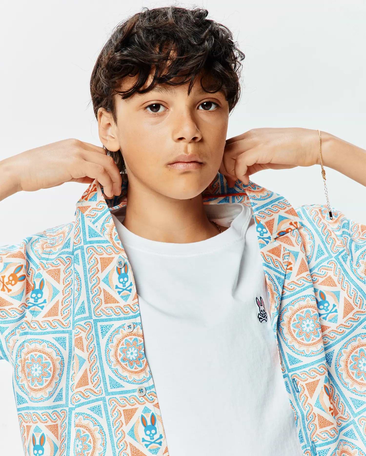 Kids Frank Tencel Shirt, a relaxed fit short-sleeve button-up in white with blue and orange mosaic bunny skull pattern, worn over a classic white jersey t-shirt with a small embroidered Psycho Bunny logo on the left chest.