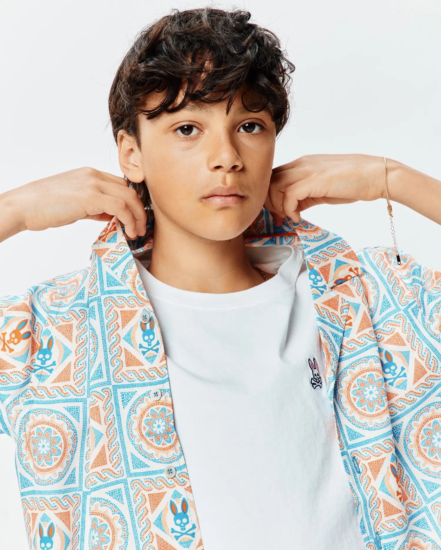 Kids Frank Tencel Shirt, a relaxed fit short-sleeve button-up in white with blue and orange mosaic bunny skull pattern, worn over a classic white jersey t-shirt with a small embroidered Psycho Bunny logo on the left chest.
