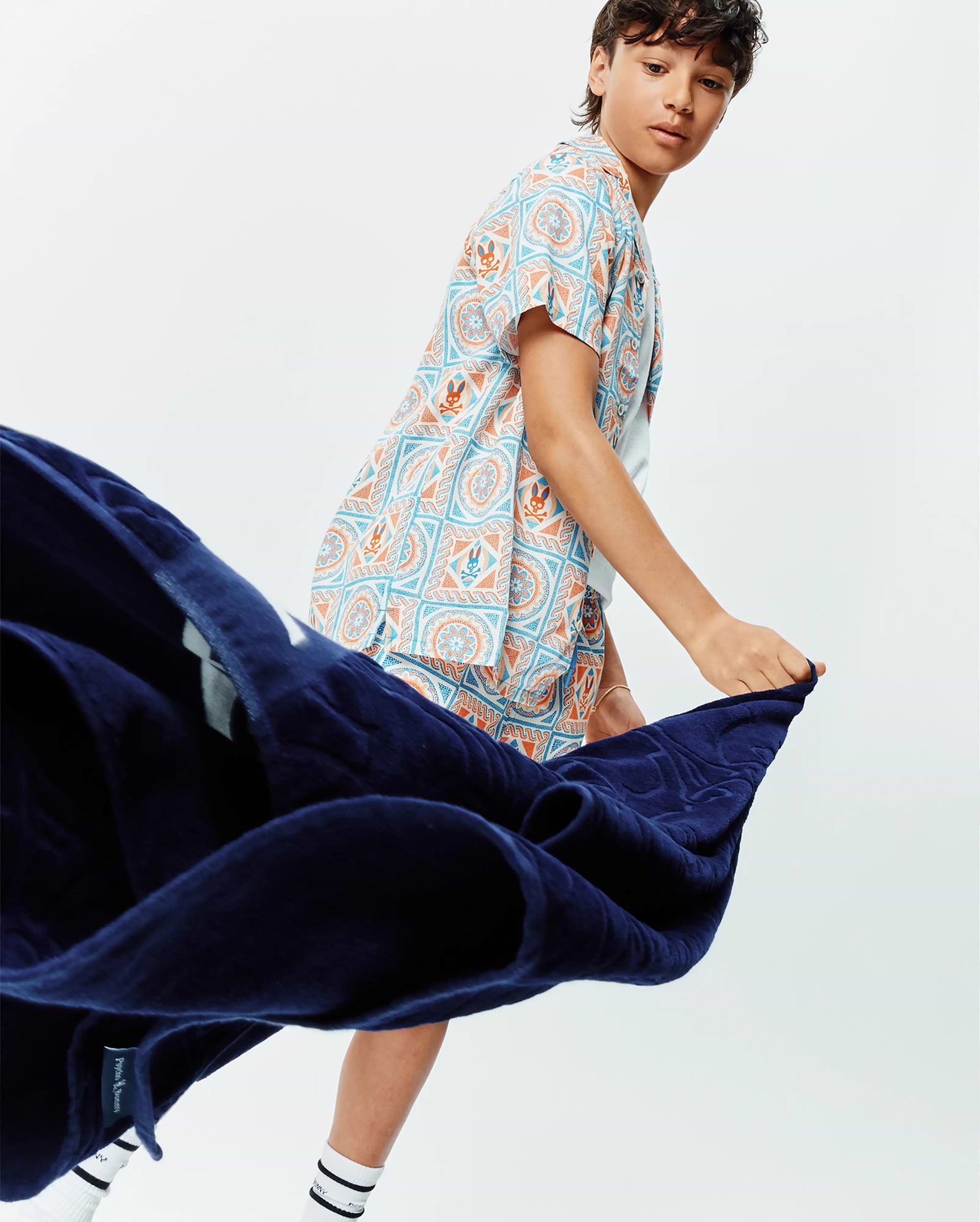 Kids Frank Tencel Shirt, a relaxed fit short-sleeve shirt featuring a vibrant all-over pattern of light blue and orange geometric motifs, mandalas, and small Psycho Bunny skull logos on a white background.