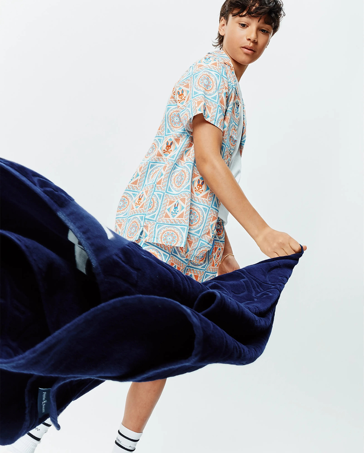 Kids Frank Tencel Shirt, a relaxed fit short-sleeve shirt featuring a vibrant all-over pattern of light blue and orange geometric motifs, mandalas, and small Psycho Bunny skull logos on a white background.