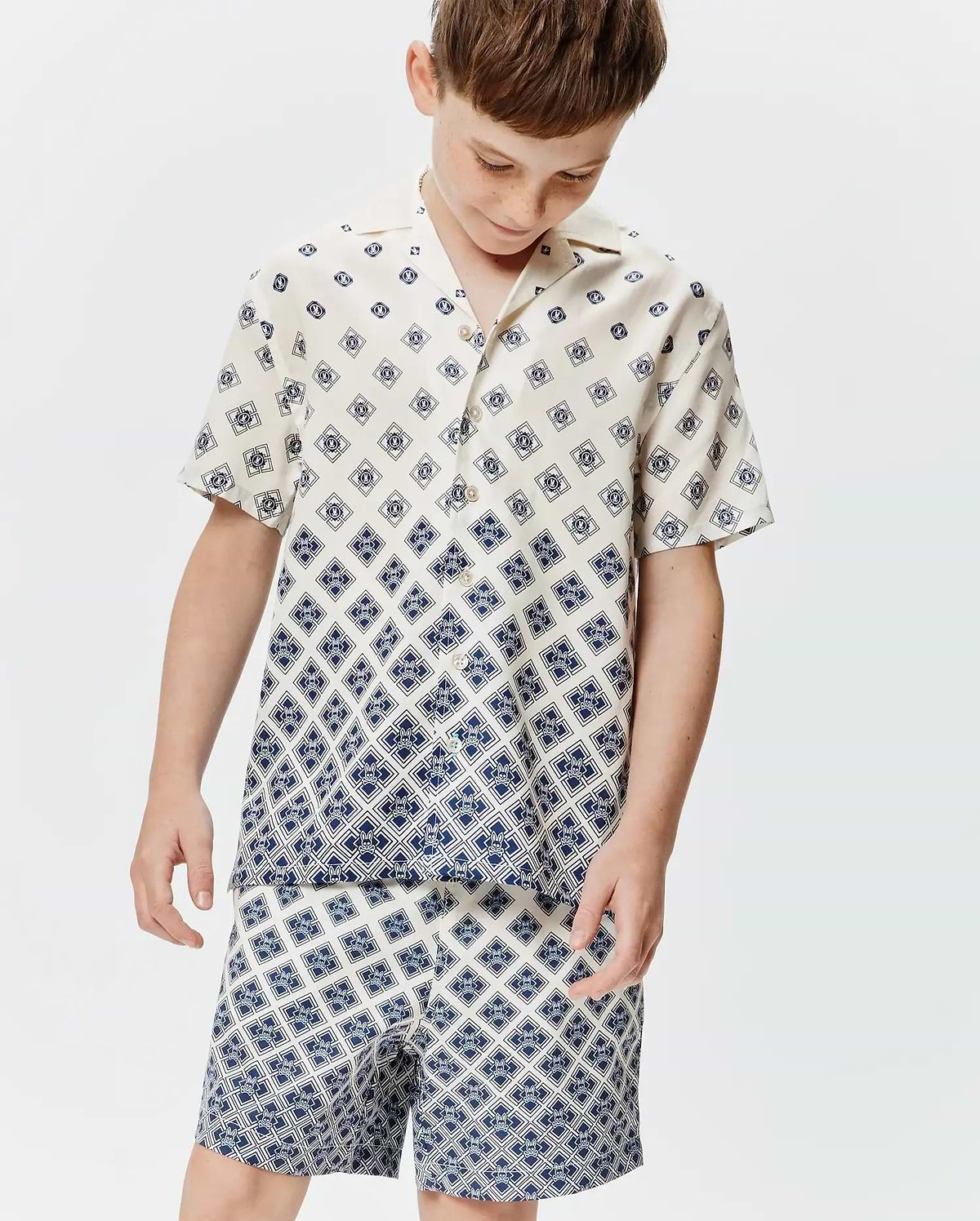 Kids Tobias Tencel Shirt, a cream short-sleeve button-up with a relaxed fit, featuring a navy blue geometric pattern of repeating Psycho Bunny skull logos, transitioning to a denser print at the hem.