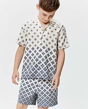 Kids Tobias Tencel Shirt, a cream short-sleeve button-up with a relaxed fit, featuring a navy blue geometric pattern of repeating Psycho Bunny skull logos, transitioning to a denser print at the hem.