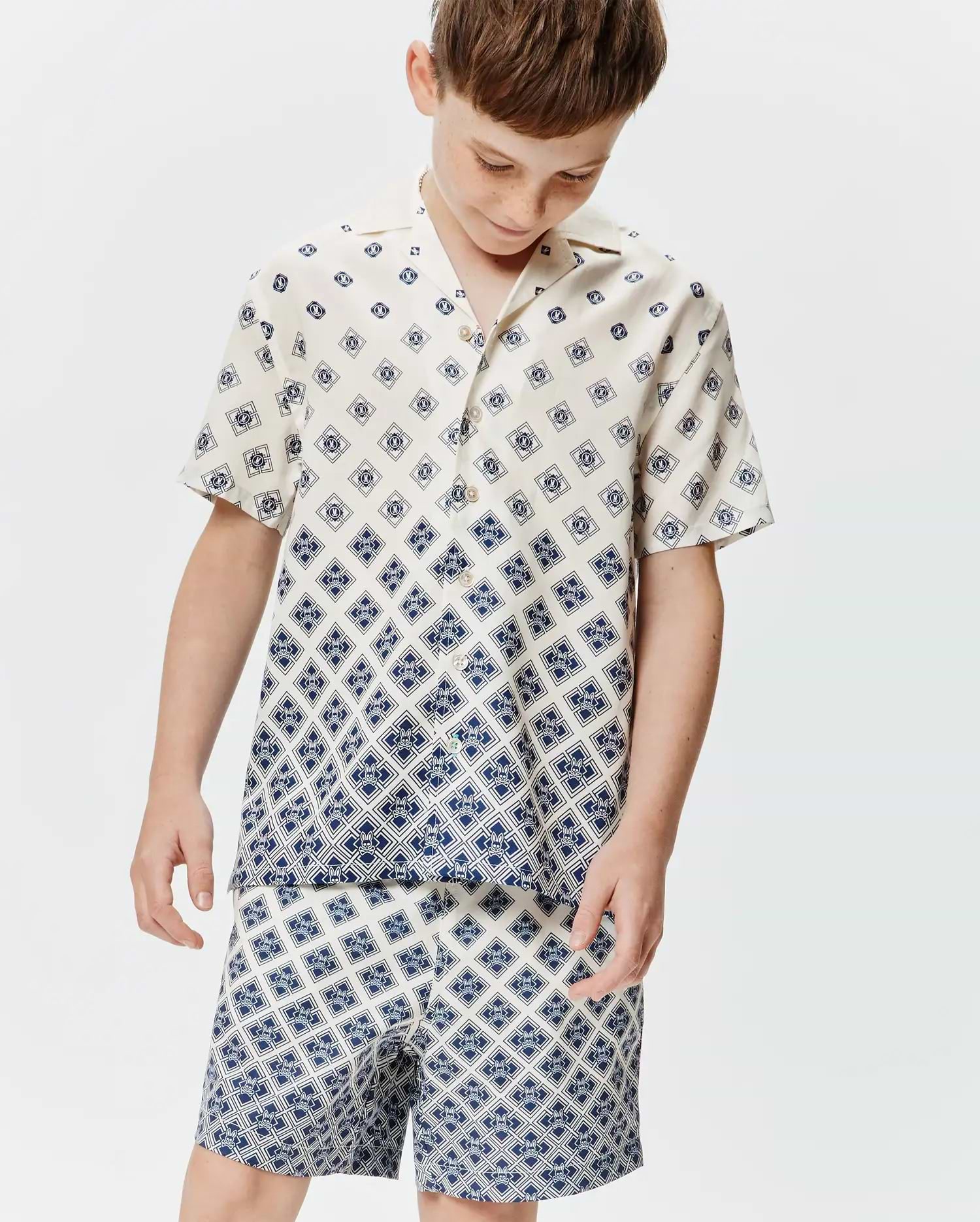 Kids Tobias Tencel Shirt, a cream short-sleeve button-up with a relaxed fit, featuring a navy blue geometric pattern of repeating Psycho Bunny skull logos, transitioning to a denser print at the hem.