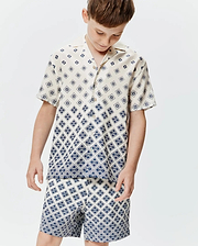 Kids Tobias Tencel Shirt, a cream short-sleeve button-up with a relaxed fit, featuring a navy blue geometric pattern of repeating Psycho Bunny skull logos, transitioning to a denser print at the hem.