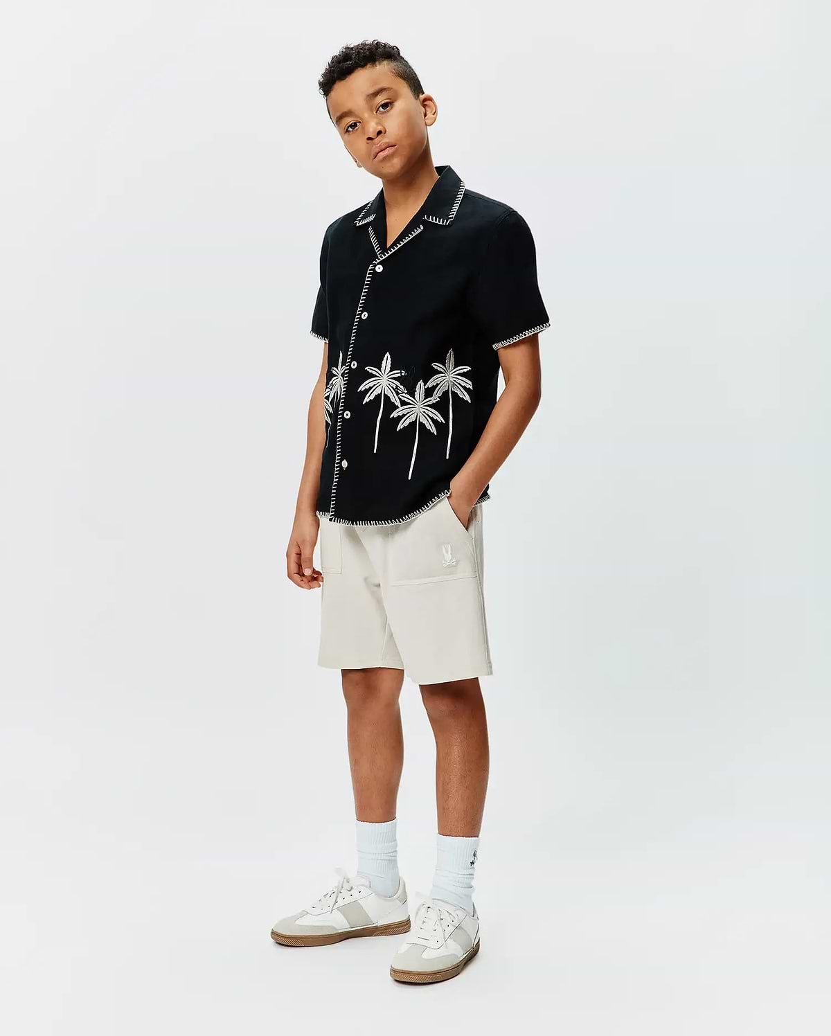 Kids Brad Linen Shirt, a black linen camp shirt with white palm tree embroidery and zig-zag stitching, paired with relaxed fit beige french terry shorts featuring a small white embroidered Psycho Bunny logo.