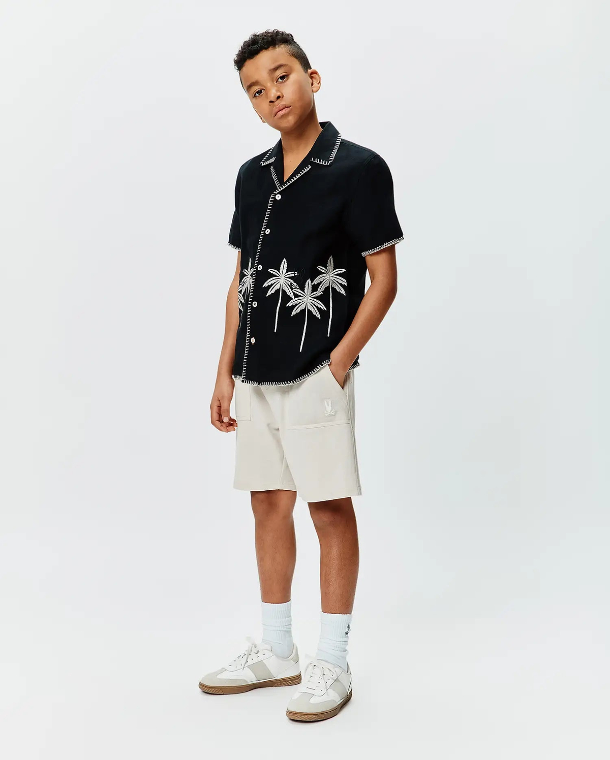 Kids Brad Linen Shirt, a black linen camp shirt with white palm tree embroidery and zig-zag stitching, paired with relaxed fit beige french terry shorts featuring a small white embroidered Psycho Bunny logo.