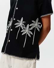 Kids Brad Linen Shirt: Black relaxed-fit linen shirt featuring white embroidered palm trees, a subtle black Psycho Bunny skull logo, and decorative white stitching along the placket, sleeves, and hem.