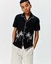 Kids Brad Linen Shirt, black with white contrast blanket stitching on collar, placket, sleeves, and hem. Features white embroidered palm trees on the front and a small orange Psycho Bunny tag on the left hem. Relaxed fit.
