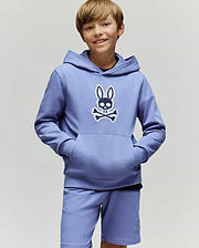 A smiling boy with light brown hair wears the Psycho Bunny KIDS RYDER EMBROIDERED BUNNY PATCH HOODIE (KT0500018), featuring an embroidered Bunny patch and crossbones logo, standing against a plain light background.
