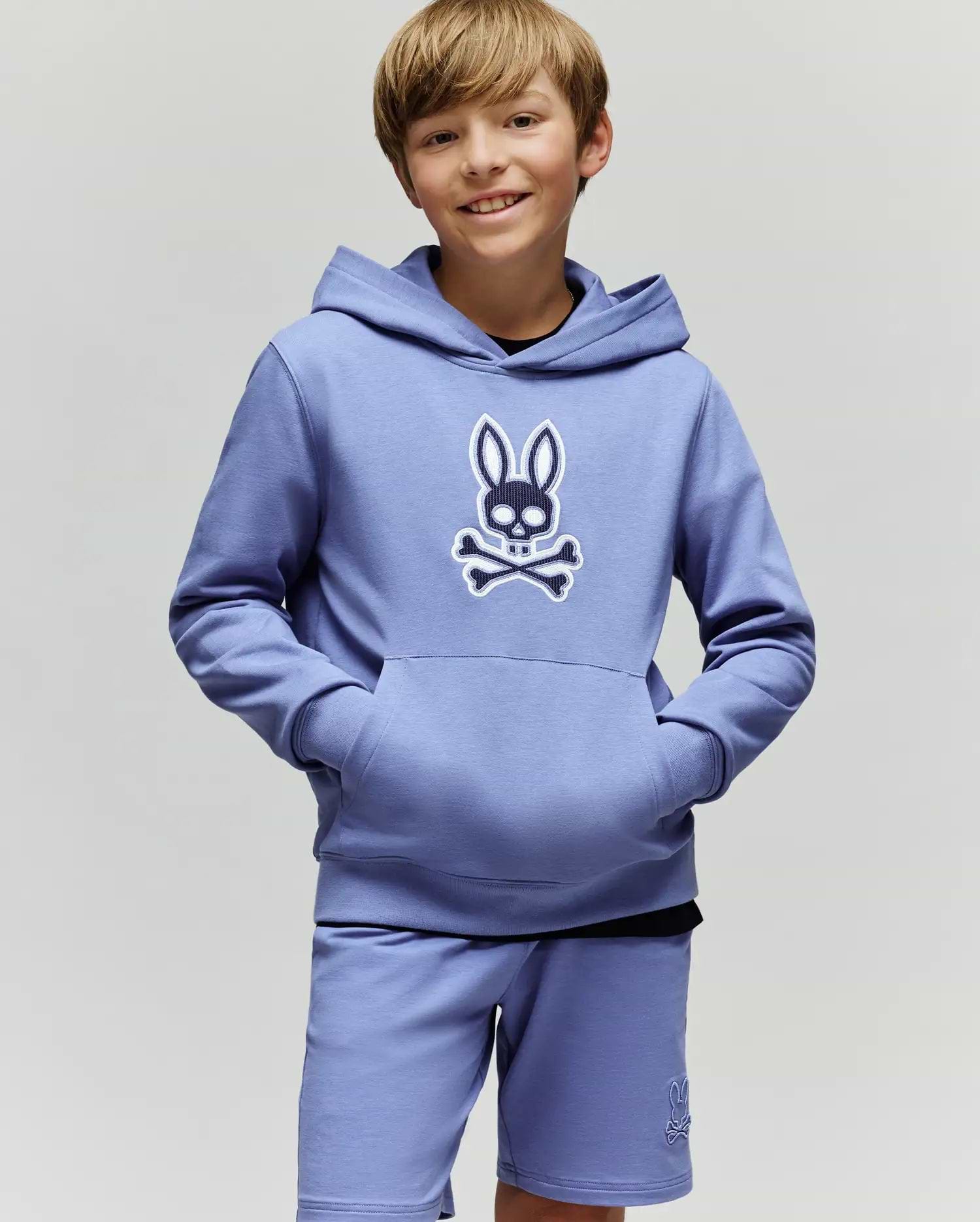 A smiling boy with light brown hair wears the Psycho Bunny KIDS RYDER EMBROIDERED BUNNY PATCH HOODIE (KT0500018), featuring an embroidered Bunny patch and crossbones logo, standing against a plain light background.