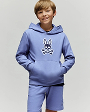 A smiling boy with light brown hair wears the Psycho Bunny KIDS RYDER EMBROIDERED BUNNY PATCH HOODIE (KT0500018), featuring an embroidered Bunny patch and crossbones logo, standing against a plain light background.