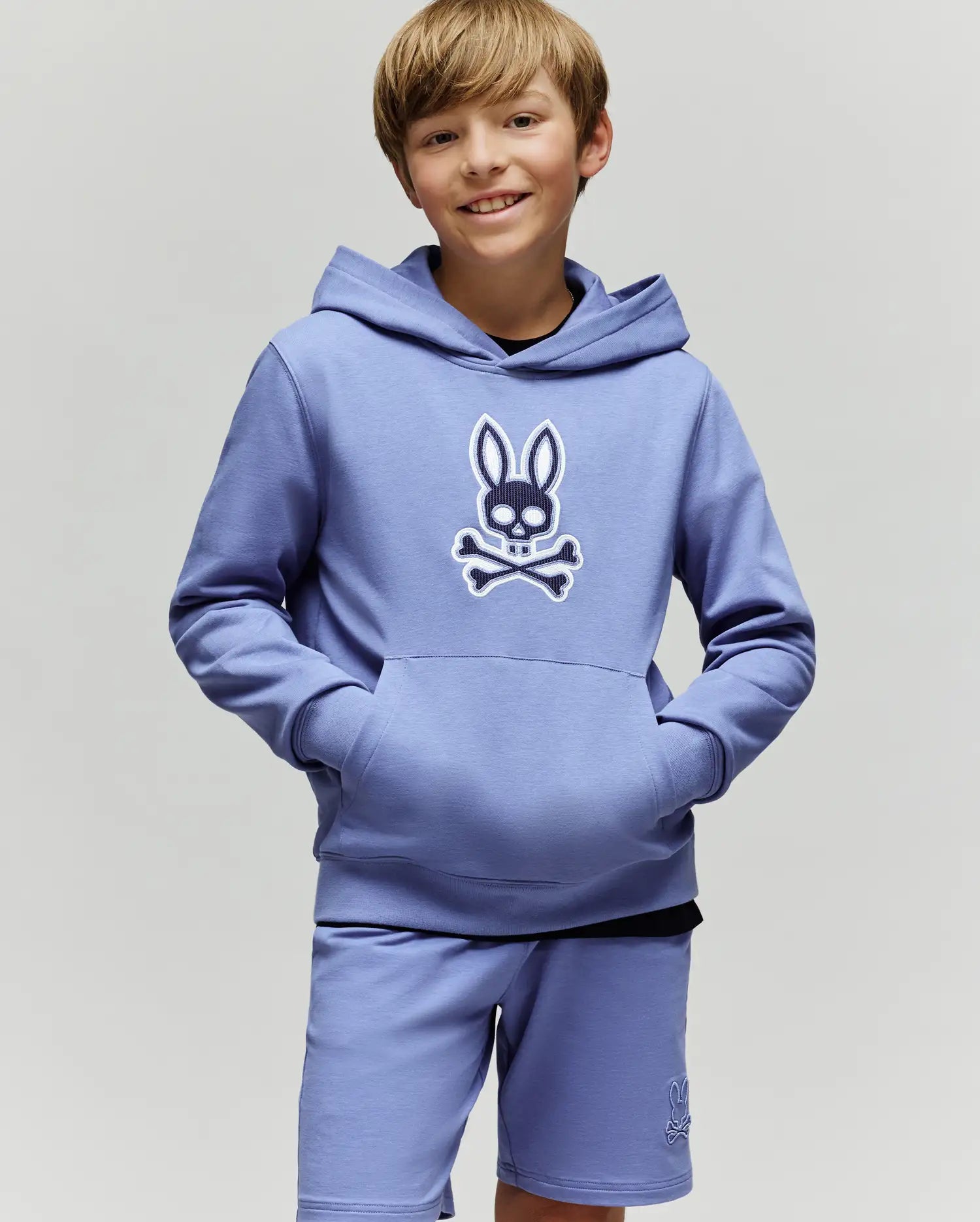 A smiling boy with light brown hair wears the Psycho Bunny KIDS RYDER EMBROIDERED BUNNY PATCH HOODIE (KT0500018), featuring an embroidered Bunny patch and crossbones logo, standing against a plain light background.