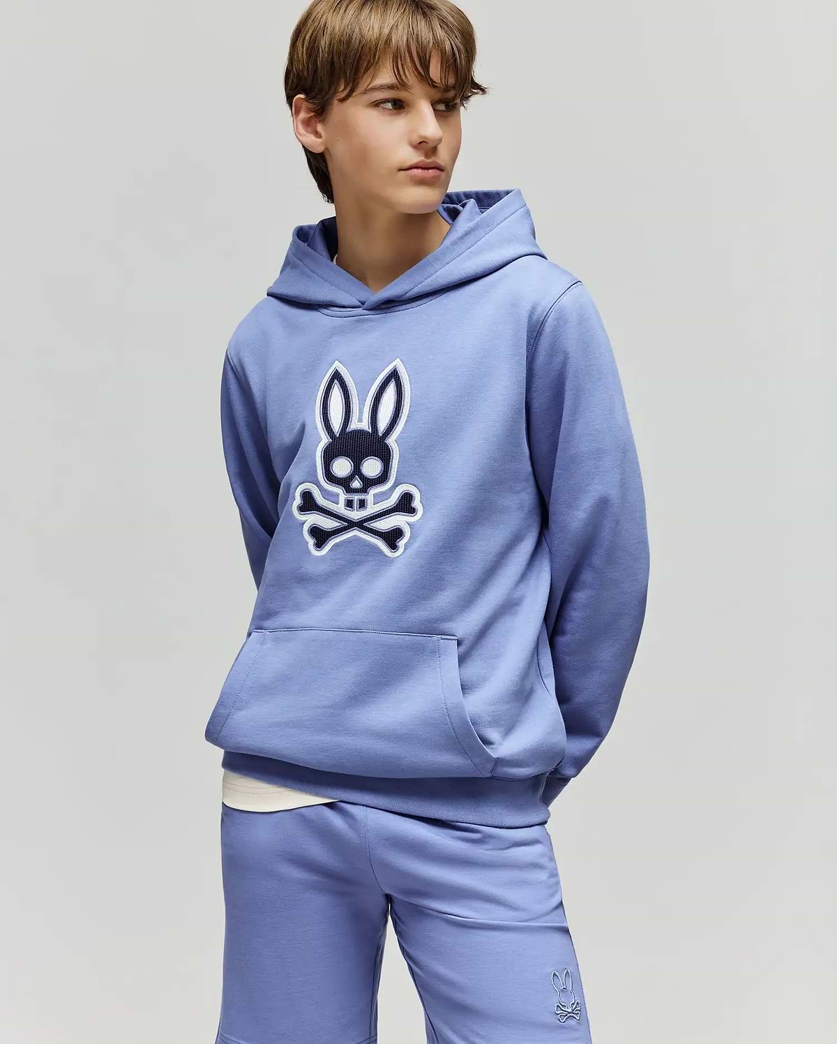 A teenage boy wears a light purple Psycho Bunny KIDS RYDER EMBROIDERED BUNNY PATCH HOODIE (KT0500018) and matching shorts, hands in pockets, gazing to the side against a plain background.