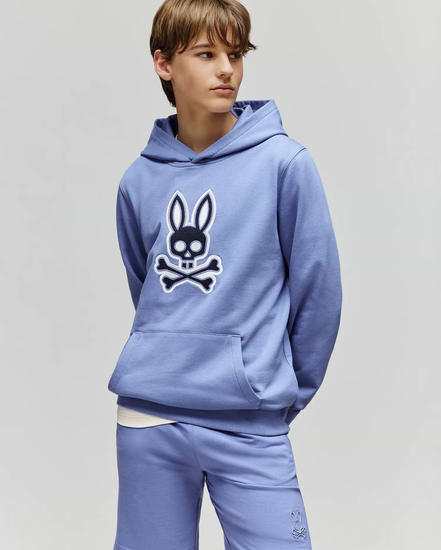 A teenage boy wears a light purple Psycho Bunny KIDS RYDER EMBROIDERED BUNNY PATCH HOODIE (KT0500018) and matching shorts, hands in pockets, gazing to the side against a plain background.