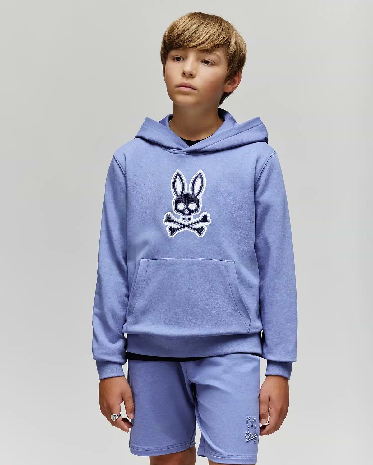 A young boy with light brown hair models the Psycho Bunny KIDS RYDER EMBROIDERED BUNNY PATCH HOODIE (KT0500018) and matching shorts, standing against a light gray background and gazing slightly to the side.