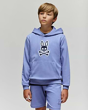 A young boy with light brown hair models the Psycho Bunny KIDS RYDER EMBROIDERED BUNNY PATCH HOODIE (KT0500018) and matching shorts, standing against a light gray background and gazing slightly to the side.
