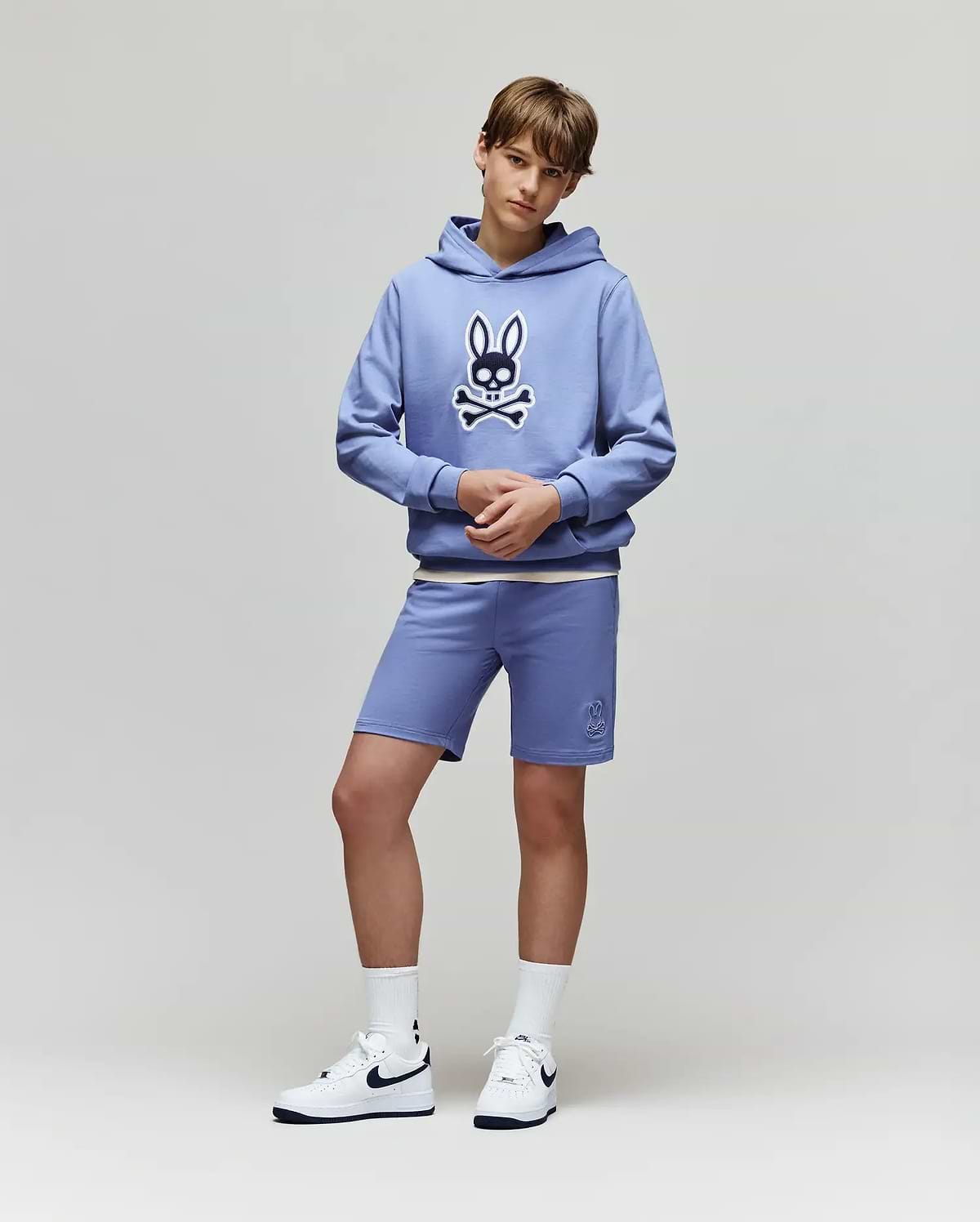 Teen boy stands against a plain background in a light blue Psycho Bunny KIDS RYDER EMBROIDERED BUNNY PATCH HOODIE (KT0500018), matching blue loungewear shorts, white socks, and white Nike sneakers.