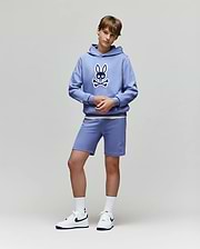 Teen boy stands against a plain background in a light blue Psycho Bunny KIDS RYDER EMBROIDERED BUNNY PATCH HOODIE (KT0500018), matching blue loungewear shorts, white socks, and white Nike sneakers.