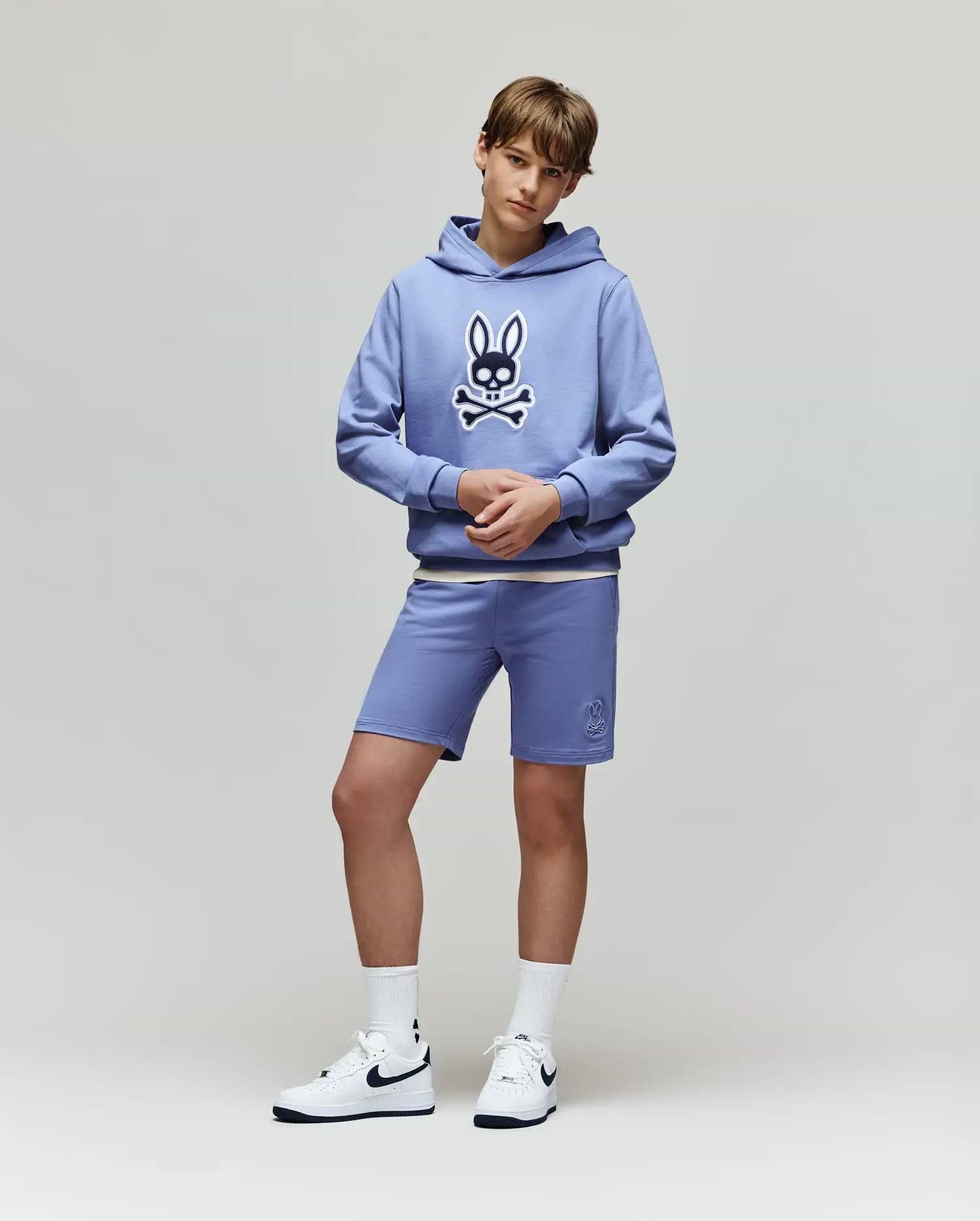 Teen boy stands against a plain background in a light blue Psycho Bunny KIDS RYDER EMBROIDERED BUNNY PATCH HOODIE (KT0500018), matching blue loungewear shorts, white socks, and white Nike sneakers.