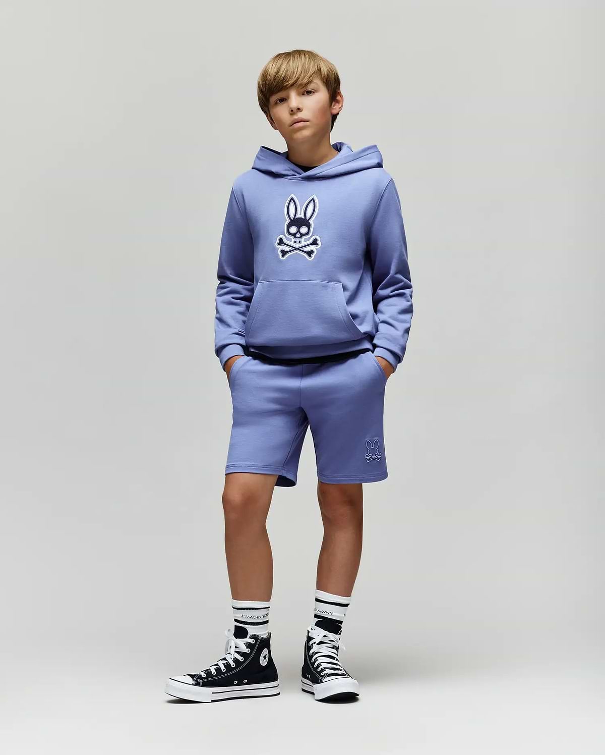 A boy stands with hands in pockets, wearing the Psycho Bunny KIDS RYDER EMBROIDERED BUNNY PATCH HOODIE (KT0500018) in lavender—paired with matching shorts, black high-top sneakers, and striped socks against a light gray background.
