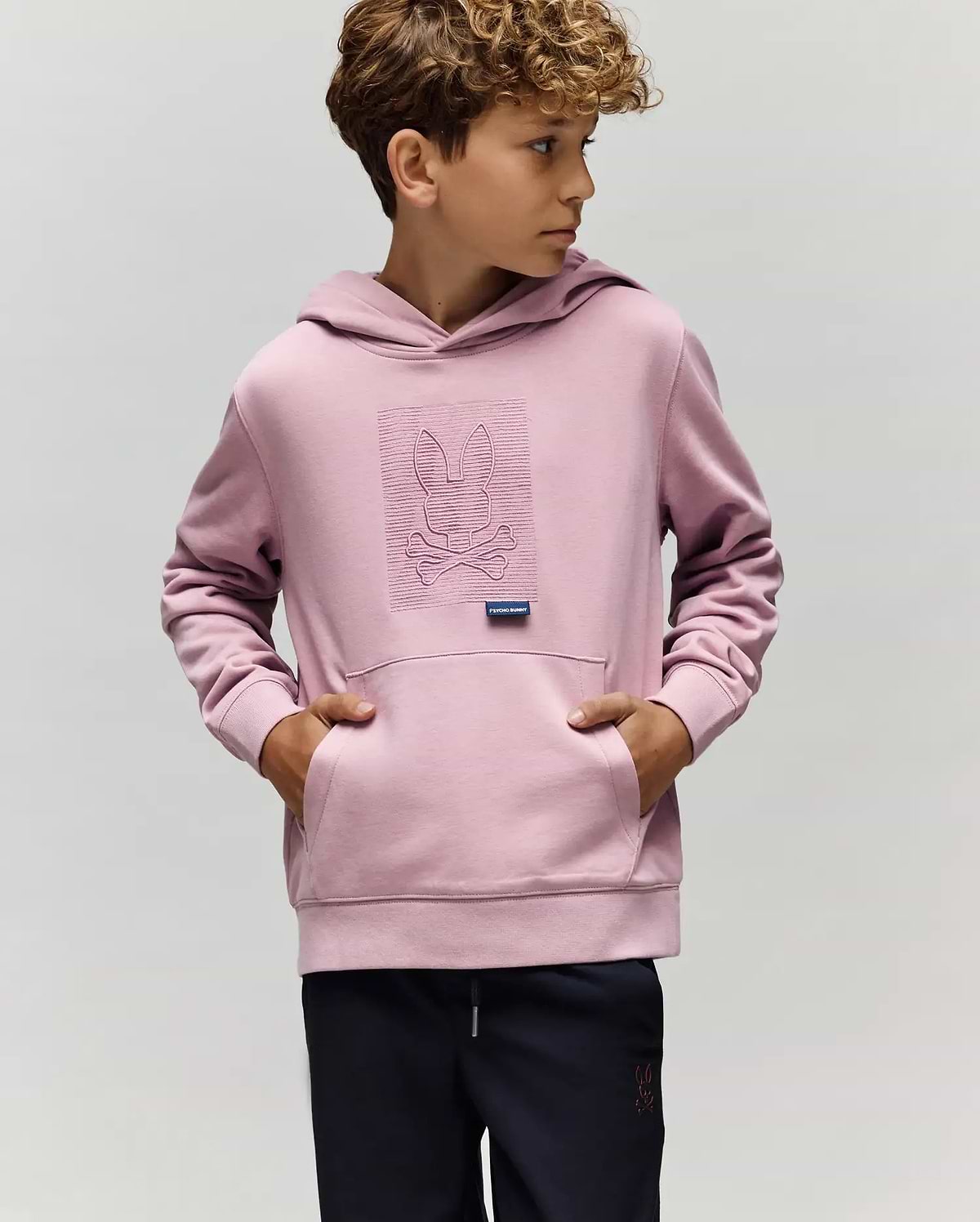 A young boy wears the KIDS GRANT HOODIE - KT0500020 by Psycho Bunny in light purple, made from a premium cotton blend with an embroidered Bunny skull and crossbones, standing with hands in the pocket against a plain background.