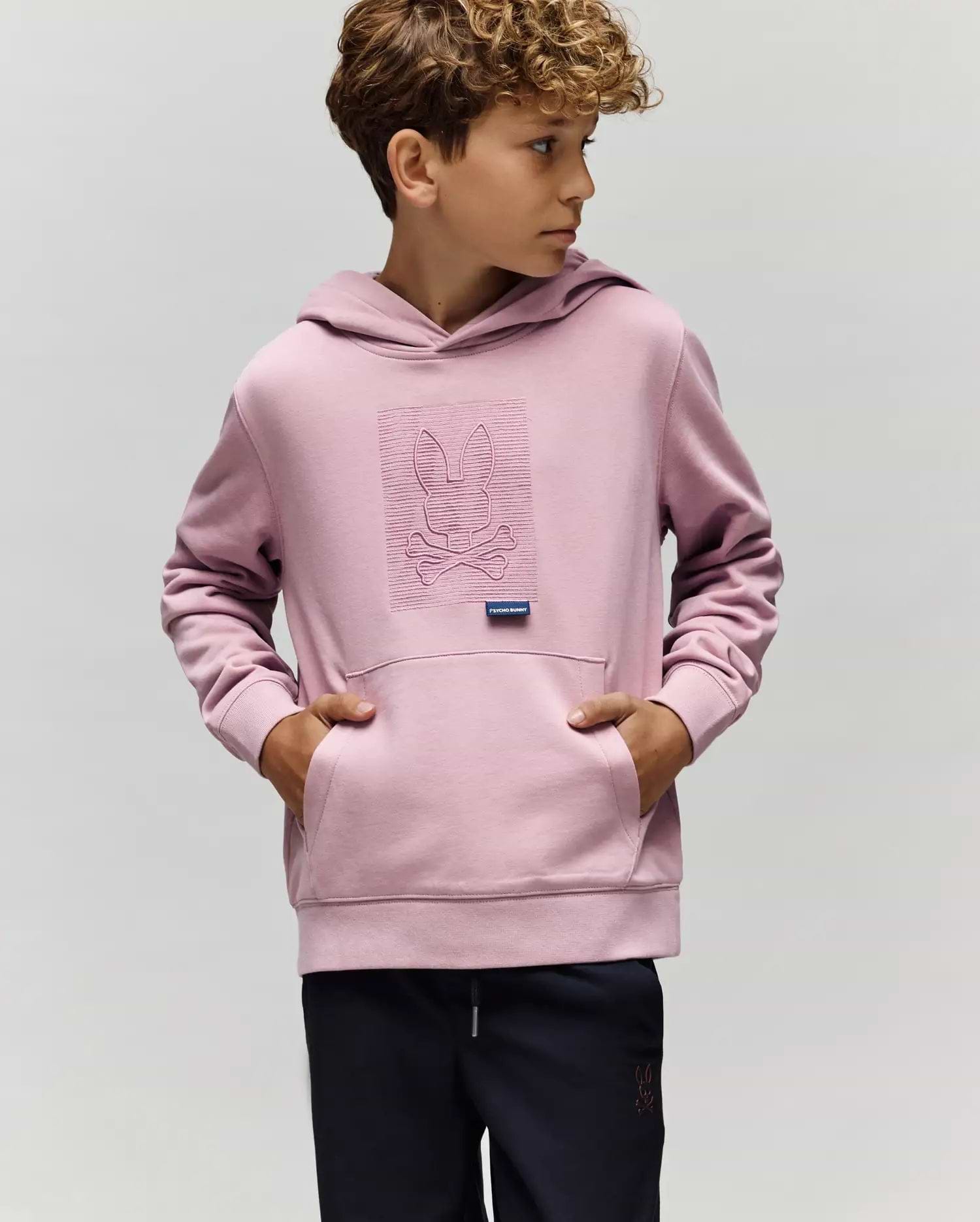 A young boy wears the KIDS GRANT HOODIE - KT0500020 by Psycho Bunny in light purple, made from a premium cotton blend with an embroidered Bunny skull and crossbones, standing with hands in the pocket against a plain background.