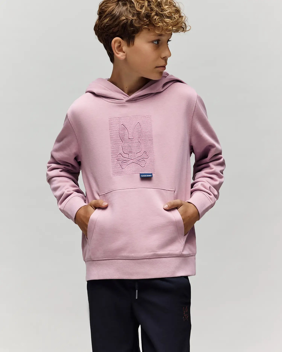A young boy wears the KIDS GRANT HOODIE - KT0500020 by Psycho Bunny in light purple, made from a premium cotton blend with an embroidered Bunny skull and crossbones, standing with hands in the pocket against a plain background.